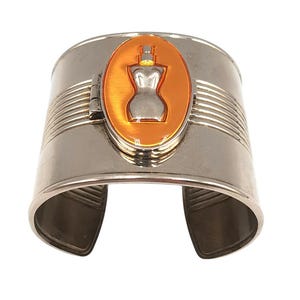 Puede incluir: Brazalete plateado con un centro ovalado naranja y plateado. El centro presenta un diseño de frasco de perfume. El brazalete tiene líneas grabadas horizontales y una abertura curva.