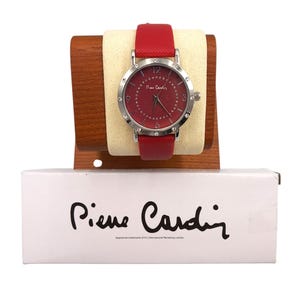 Può includere: Un orologio da polso Pierre Cardin rosso con cassa color argento e cinturino in pelle rosso abbinato. Il quadrante dell'orologio è rosso intenso con lancette e indici argentati. L'orologio è esposto su un supporto in legno, con il logo Pierre Cardin sulla scatola.