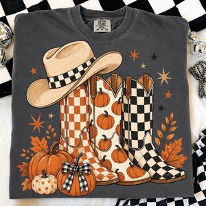 May include: A charcoal gray crewneck sweatshirt featuring a graphic print of a beige cowboy hat with a checkered band, three cowboy boots with orange checkered, pumpkin, and black checkered patterns, pumpkins, and orange autumn leaves.