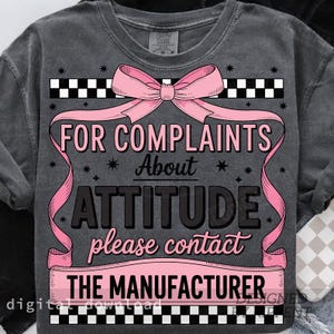 May include: Dark gray sweatshirt with the text "FOR COMPLAINTS About ATTITUDE please contact THE MANUFACTURER" in pink and black. The design includes a pink bow and ribbon, and a checkered pattern.