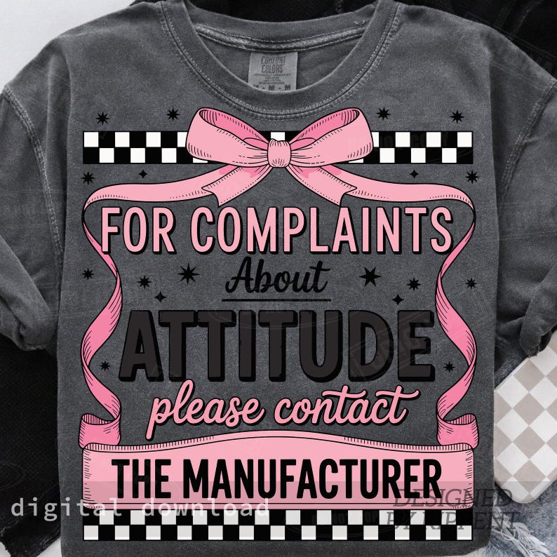 May include: Dark gray sweatshirt with the text "FOR COMPLAINTS About ATTITUDE please contact THE MANUFACTURER" in pink and black. The design includes a pink bow and ribbon, and a checkered pattern.