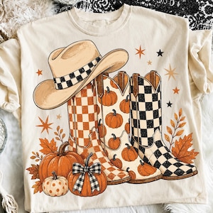 May include: A cream-colored t-shirt featuring a graphic print of a tan cowboy hat with a checkered band, three western-style boots with orange checkered, pumpkin, and black checkered patterns, orange pumpkins, and scattered star and leaf shapes.