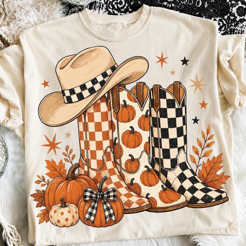 May include: A cream-colored t-shirt featuring a graphic print of a tan cowboy hat with a checkered band, three western-style boots with orange checkered, pumpkin, and black checkered patterns, orange pumpkins, and scattered star and leaf shapes.