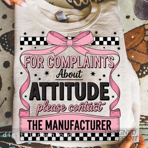 May include: A beige sweatshirt with the text "FOR COMPLAINTS About ATTITUDE please contact THE MANUFACTURER" in black and pink. The design includes a pink bow, ribbons, and star accents. The top has a crew neck.