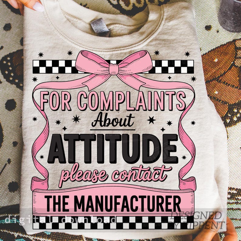 May include: A beige sweatshirt with the text "FOR COMPLAINTS About ATTITUDE please contact THE MANUFACTURER" in black and pink. The design includes a pink bow, ribbons, and star accents. The top has a crew neck.