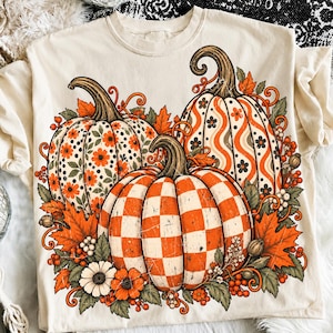 Könnte beinhalten: Ein cremefarbenes T-Shirt mit einem Grafikdruck von drei Kürbissen. Ein Kürbis hat ein Blumenmuster, einer ein Wellenlinien- und Blumenmuster und der dritte ein Karomuster. Das Design enthält orangefarbene Blätter und Beeren.
