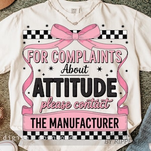 May include: Cream-colored t-shirt with a pink bow and ribbon design. The text reads "FOR COMPLAINTS ABOUT ATTITUDE please contact THE MANUFACTURER". Black and white checkered patterns frame the text.