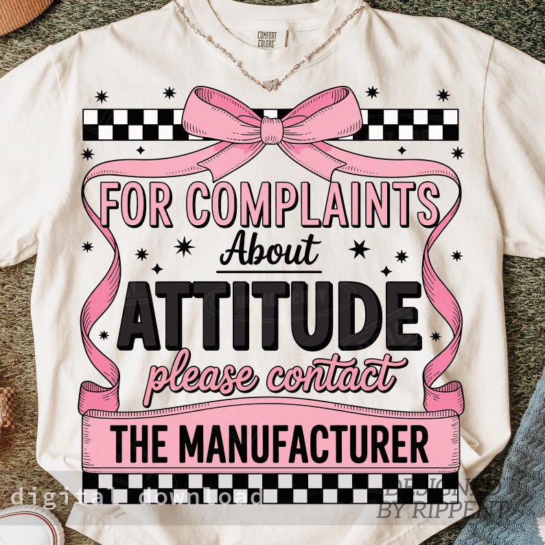 May include: Cream-colored t-shirt with a pink bow and ribbon design. The text reads "FOR COMPLAINTS ABOUT ATTITUDE please contact THE MANUFACTURER". Black and white checkered patterns frame the text.