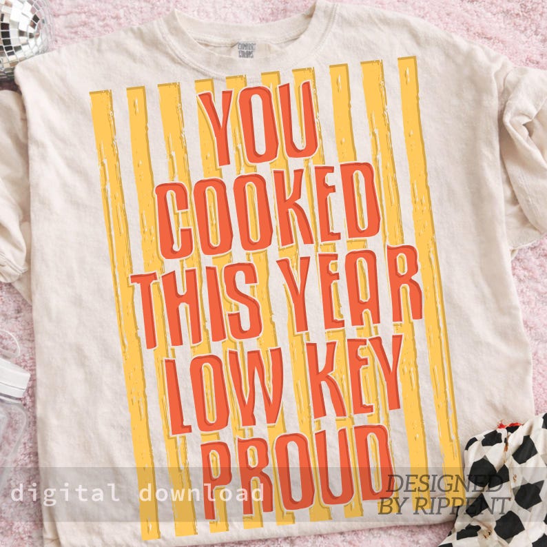 May include: Cream-colored t-shirt with orange text that reads "YOU COOKED THIS YEAR LOW KEY PROUD" with yellow vertical stripes. The words "digital download" and "DESIGNED BY RIPPNT" are also visible.