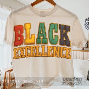 May include: Cream-colored t-shirt with the words "BLACK EXCELLENCE" in bold, colorful letters. The word "BLACK" is in red, yellow, green, and black. The shirt is hanging on a wooden hanger.
