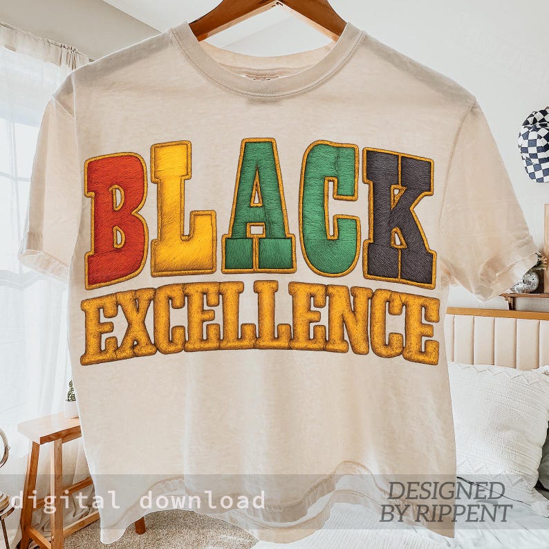 May include: Cream-colored t-shirt with the words "BLACK EXCELLENCE" in bold, colorful letters. The word "BLACK" is in red, yellow, green, and black. The shirt is hanging on a wooden hanger.