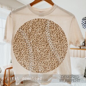 May include: Cream-colored t-shirt featuring a large baseball design with a leopard print pattern. The baseball design has white stitching details. The words "digital download" and "DESIGNED BY RIPPENT" are printed at the bottom of the shirt.