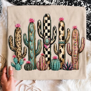 May include: A beige t-shirt featuring a graphic print of various cacti. The cacti display diverse patterns including leopard print, black and white checkerboard, and cow print, accented with turquoise stones, pink flowers, and geometric designs.