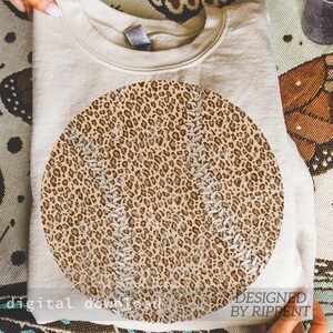 May include: A beige sweatshirt with a large baseball design. The baseball graphic features a leopard print pattern with white stitching details. The words "digital download" and "designed by Rippent" are printed on the bottom of the image.