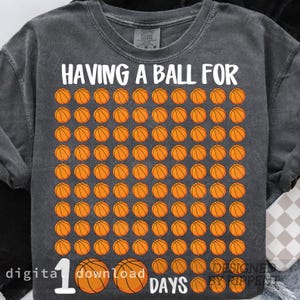 May include: Dark gray t-shirt with the text "HAVING A BALL FOR 100 DAYS" in white. The shirt features a grid of orange basketballs. The design is centered on the front of the shirt.