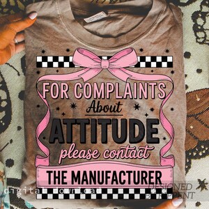 May include: A brown t-shirt with a pink bow and the text "FOR COMPLAINTS About ATTITUDE please contact THE MANUFACTURER". The design includes black and white checkered patterns and pink ribbons.