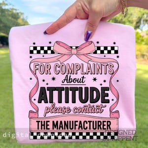 May include: A light pink sweatshirt with the text "FOR COMPLAINTS About ATTITUDE please contact THE MANUFACTURER". The design includes a pink bow, ribbons, and black and white checkered borders. The words are in black and pink.