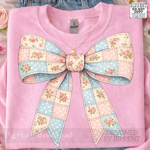 May include: A light pink sweatshirt featuring a large bow design. The bow is a patchwork of pink and blue squares with floral patterns and polka dots. A gold heart pendant necklace hangs from the neckline. The text "RIPPENT DO NOT COPY ORIGINAL DESIGN" is visible.