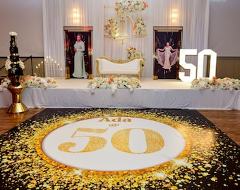 Pode incluir: Um palco branco com um trono dourado e branco, duas fotos emolduradas de uma mulher e um grande número 50 iluminado. O palco é decorado com flores brancas e pêssego. Uma pista de dança preta e dourada com o texto "Ada @ 50" está em primeiro plano.