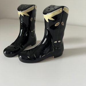 May include: A pair of black ceramic salt and pepper shakers shaped like cowboy boots. The boots have a glossy finish with tan and yellow accents on the tops. Each boot has small holes on the top for dispensing salt and pepper.