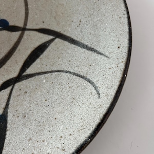 May include: A handmade ceramic plate with a speckled, off-white surface. The plate features a dark brown rim and is decorated with black, stylized floral designs and small blue accents. The plate's design is simple and elegant.