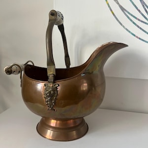 May include: A copper-toned metal coal scuttle with a decorative lion's head emblem. The scuttle has a curved spout and a handle with ceramic accents. The base is round and raised, and the handle is arched.