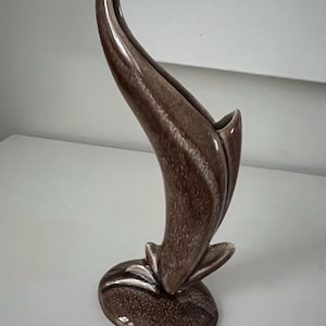 May include: A brown ceramic vase with a unique, curved design. The vase has a flared opening and a textured surface, with a base that resembles a flower. The vase is approximately 10 inches tall.