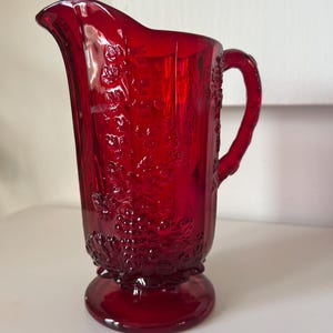 May include: A ruby red glass pitcher with a decorative floral pattern. The pitcher has a handle and a flared spout. The base is round and the pitcher is made of a transparent glass material. The pitcher is a vintage style and is suitable for serving beverages.
