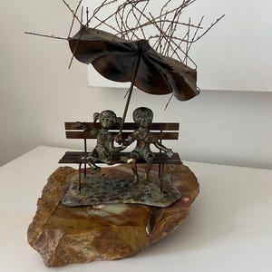 May include: A bronze sculpture of two children sitting on a bench under an umbrella. The sculpture is mounted on a natural stone base. The umbrella has delicate wire details, and the children are holding hands.