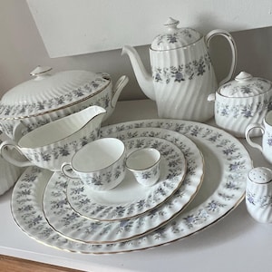 May include: A white porcelain tea set with a blue floral pattern and gold trim. The set includes a teapot, sugar bowl, creamer, serving dishes, plates, cups, and salt and pepper shakers. The dishes have a ribbed design.