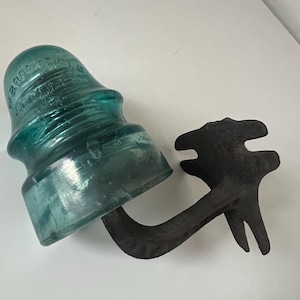 May include: A vintage teal glass insulator with the text "BROOKFIELD" and a black metal bracket. The insulator is a classic design, likely used for electrical wiring. The bracket is a dark, weathered metal.