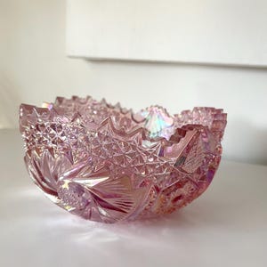 May include: A pink iridescent glass bowl with a scalloped edge and a detailed, geometric pattern. The bowl is made of pressed glass and has a vintage look.