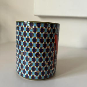 Puede incluir: Taza cil&iacute;ndrica de cer&aacute;mica con un patr&oacute;n geom&eacute;trico en azul, morado y blanco. La taza presenta detalles de abejas doradas y un dise&ntilde;o rojo en el lateral. El borde es verde.
