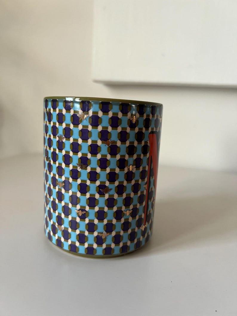 Puede incluir: Taza cil&iacute;ndrica de cer&aacute;mica con un patr&oacute;n geom&eacute;trico en azul, morado y blanco. La taza presenta detalles de abejas doradas y un dise&ntilde;o rojo en el lateral. El borde es verde.