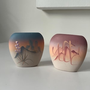 May include: Two small ceramic vases with hand-painted desert scenes. One vase features a purple flower and a mountain range in shades of orange, blue, and brown. The other vase features a desert landscape with tall, thin plants in shades of orange, pink, and brown.