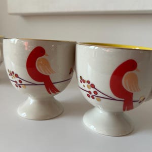May include: Two white ceramic egg cups with a gold rim and a red bird design. The birds are perched on a branch with gold berries.