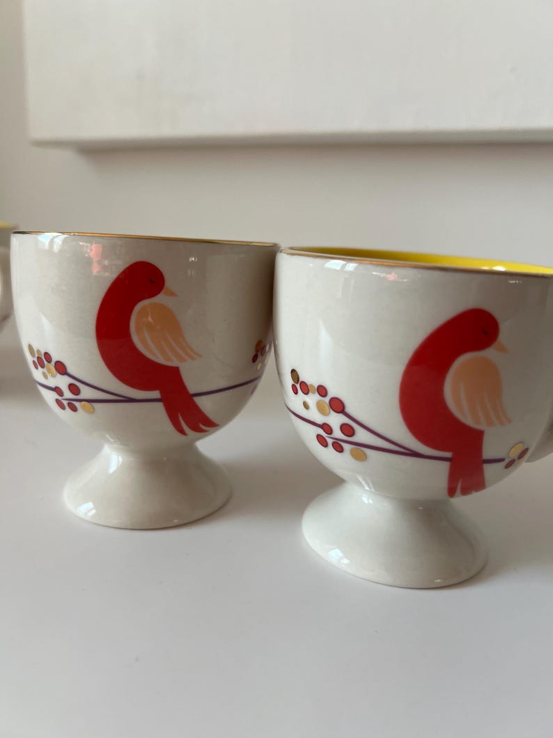 May include: Two white ceramic egg cups with a gold rim and a red bird design. The birds are perched on a branch with gold berries.