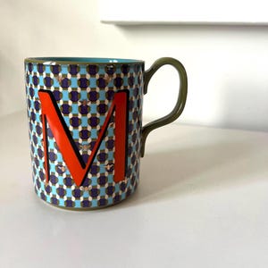 Puede incluir: Una taza de cer&aacute;mica con un patr&oacute;n geom&eacute;trico azul, morado y dorado. La taza presenta una gran letra "M" naranja y un asa verde. Peque&ntilde;as ilustraciones de abejas doradas est&aacute;n dispersas por el dise&ntilde;o. El interior es turquesa.