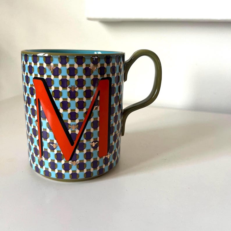 Puede incluir: Una taza de cer&aacute;mica con un patr&oacute;n geom&eacute;trico azul, morado y dorado. La taza presenta una gran letra "M" naranja y un asa verde. Peque&ntilde;as ilustraciones de abejas doradas est&aacute;n dispersas por el dise&ntilde;o. El interior es turquesa.