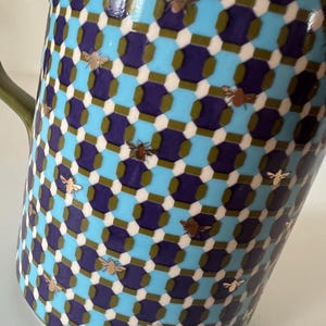 Puede incluir: Una taza de cer&aacute;mica con un patr&oacute;n geom&eacute;trico en verde azulado, azul marino y blanco. La taza presenta ilustraciones de abejas doradas dispersas por la superficie. El asa es de color verde liso.