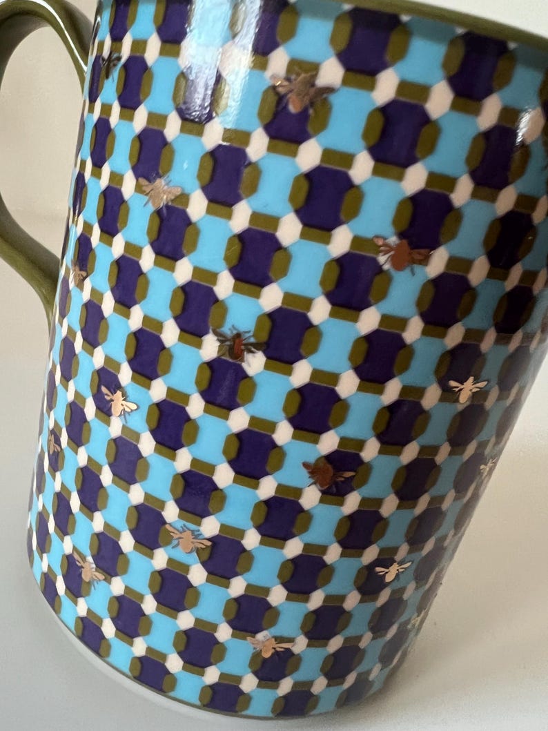 Puede incluir: Una taza de cer&aacute;mica con un patr&oacute;n geom&eacute;trico en verde azulado, azul marino y blanco. La taza presenta ilustraciones de abejas doradas dispersas por la superficie. El asa es de color verde liso.