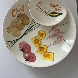 May include: A cream-colored ceramic chip and dip serving set. The large plate has illustrations of chips, tomatoes, and shrimp. The small dip bowl has the word "dip" and a leaf design. The word "chip" is on the large plate.