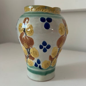 May include: A small, white ceramic vase with a green and yellow rim. The vase is decorated with brown, yellow, and blue floral patterns.