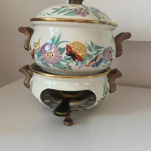 May include: A two-piece, off-white enamelware fondue set. The set includes a lidded pot with floral designs in purple, yellow, and red, and a matching base with a burner. The pot has gold trim and brown handles.