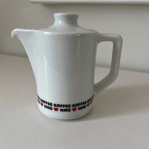 May include: White ceramic creamer with a lid and handle. The creamer has a spout for easy pouring. The base of the creamer features the words "KAFFEE HAG" in black text with red hearts. A classic design for serving coffee or cream.