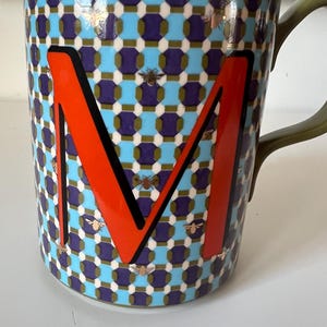 Puede incluir: Una taza de cer&aacute;mica con un patr&oacute;n geom&eacute;trico en azul, morado y blanco, con una letra "M" naranja. La taza tiene un asa verde y est&aacute; decorada con ilustraciones de abejas.