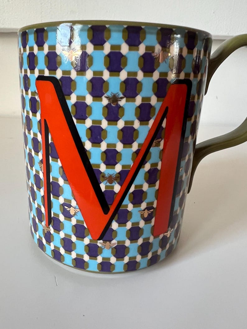 Puede incluir: Una taza de cer&aacute;mica con un patr&oacute;n geom&eacute;trico en azul, morado y blanco, con una letra "M" naranja. La taza tiene un asa verde y est&aacute; decorada con ilustraciones de abejas.