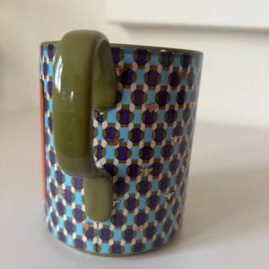 Puede incluir: Una taza de cer&aacute;mica con un patr&oacute;n geom&eacute;trico en azul, morado y blanco, acentuado con motivos de abejas doradas. El asa es verde oliva y se ve una franja naranja vertical. Un art&iacute;culo de cocina decorativo.