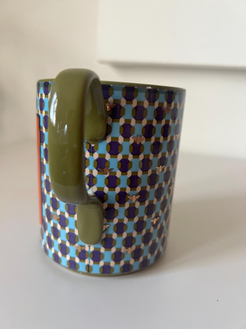 Puede incluir: Una taza de cer&aacute;mica con un patr&oacute;n geom&eacute;trico en azul, morado y blanco, acentuado con motivos de abejas doradas. El asa es verde oliva y se ve una franja naranja vertical. Un art&iacute;culo de cocina decorativo.