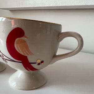 May include: A white ceramic mug with a gold rim and a red and orange bird design. The mug has a pedestal base.